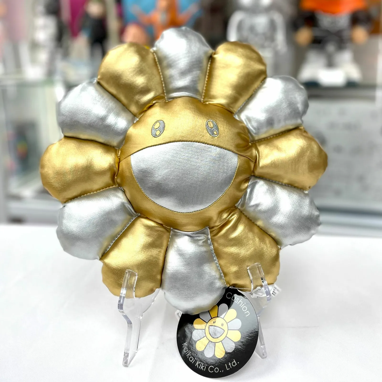 Cojín Takashi Murakami Flower Cushion en plata y oro, mostrando una cara sonriente.