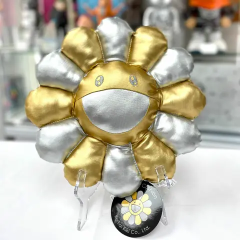 Cojín Takashi Murakami Flower Cushion en plata y oro, mostrando una cara sonriente.