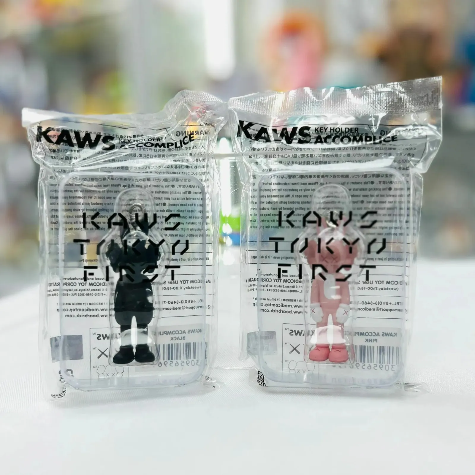 Ensemble de deux porte-clés Kaws Accomplice, l'un rose et l'autre noir, posés sur une surface claire.
